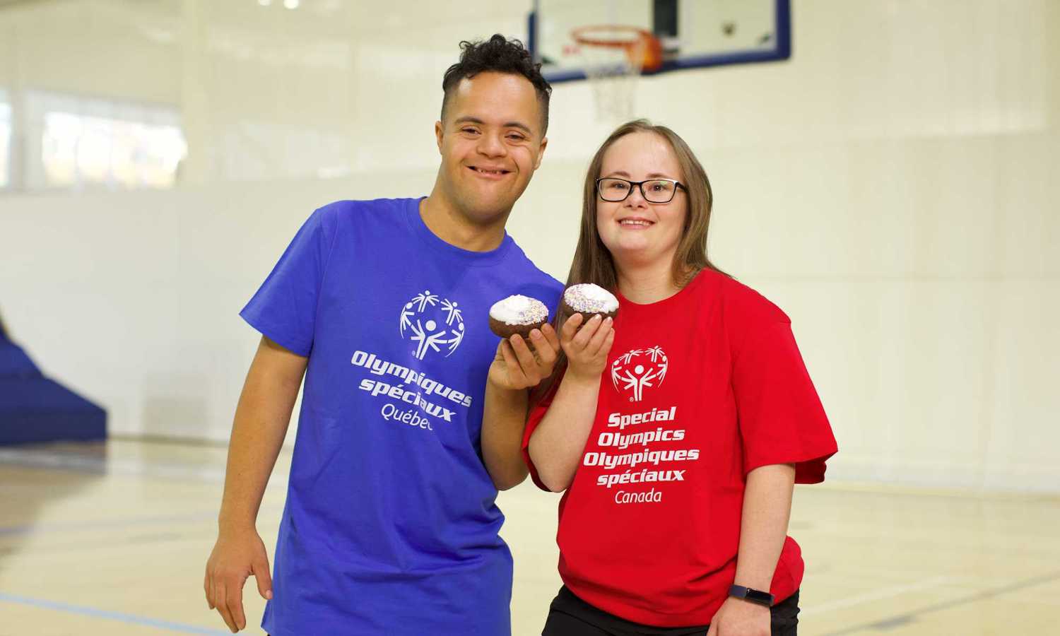 Two athletes smiling at the camera. The male on the left is wearing a blue Special Olympics Quebec t-shirt and the woman on the first is wearing a red Special Olympics Canada tshirt. Both are smiling and holding a donut. 