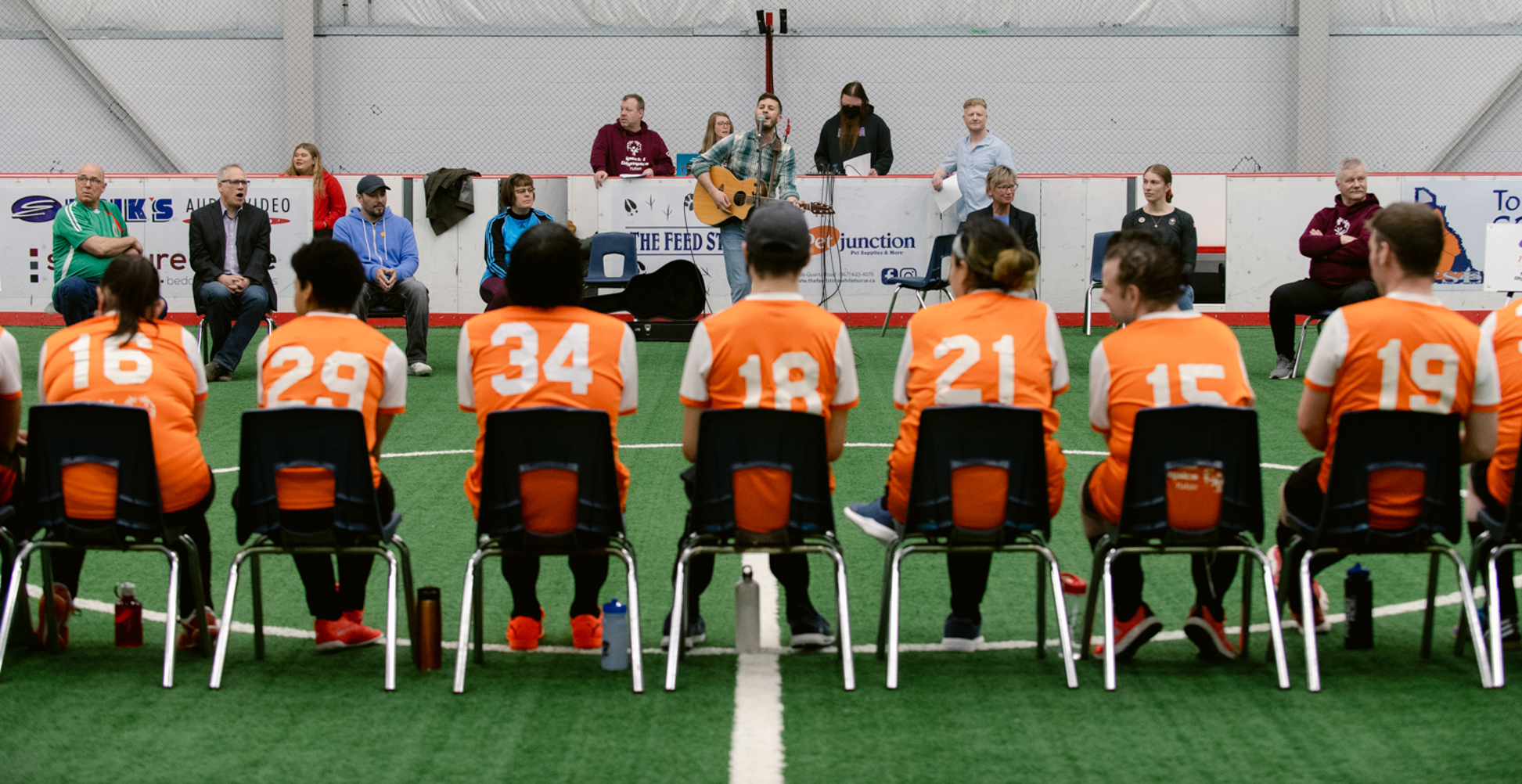 A soccer team is sitting on chairs with their backs turned to the camera. They are watching a performance of someone singing. They are wearing orange jerseys with white numbers on the back.