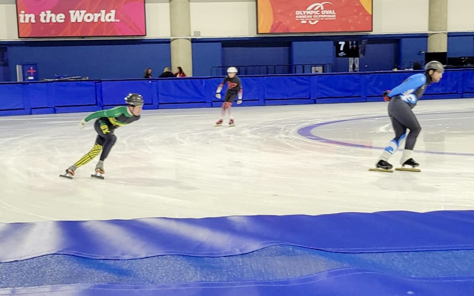 An action shot photo of speed skaters on the ice. Three speed skaters can be seen going around the edge of the rink. They are slightly out of focus due to the speed they are skating.