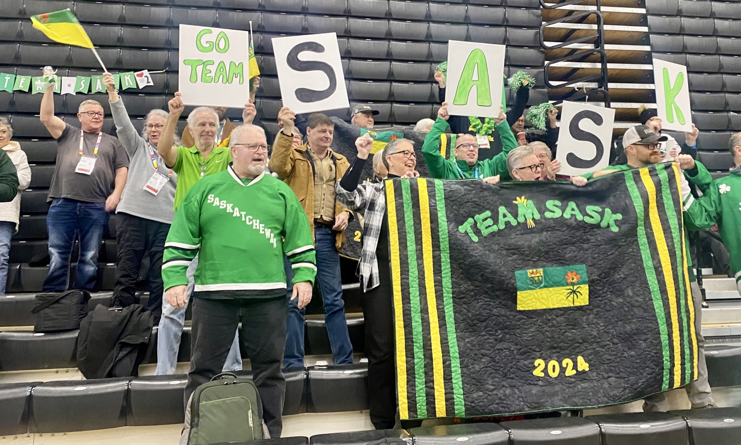 A crowd of people in green, are sitting on the bleachers cheering for Team Saskatchewan. They are holding up flags, banners, and poster boards showing support for the team. Everyone looks happy and are cheering the athletes on.