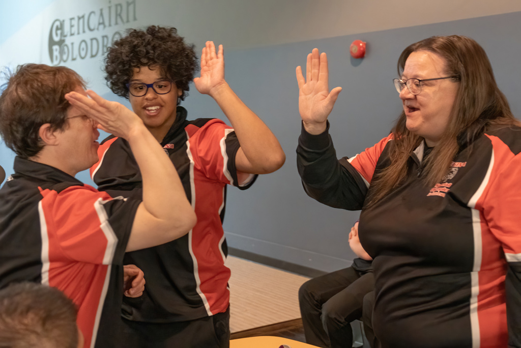 Three bowling players are smiling and giving high-fives to each other. They are all wearing the same black and red bowling shirt. They are standing in a bowling alley.