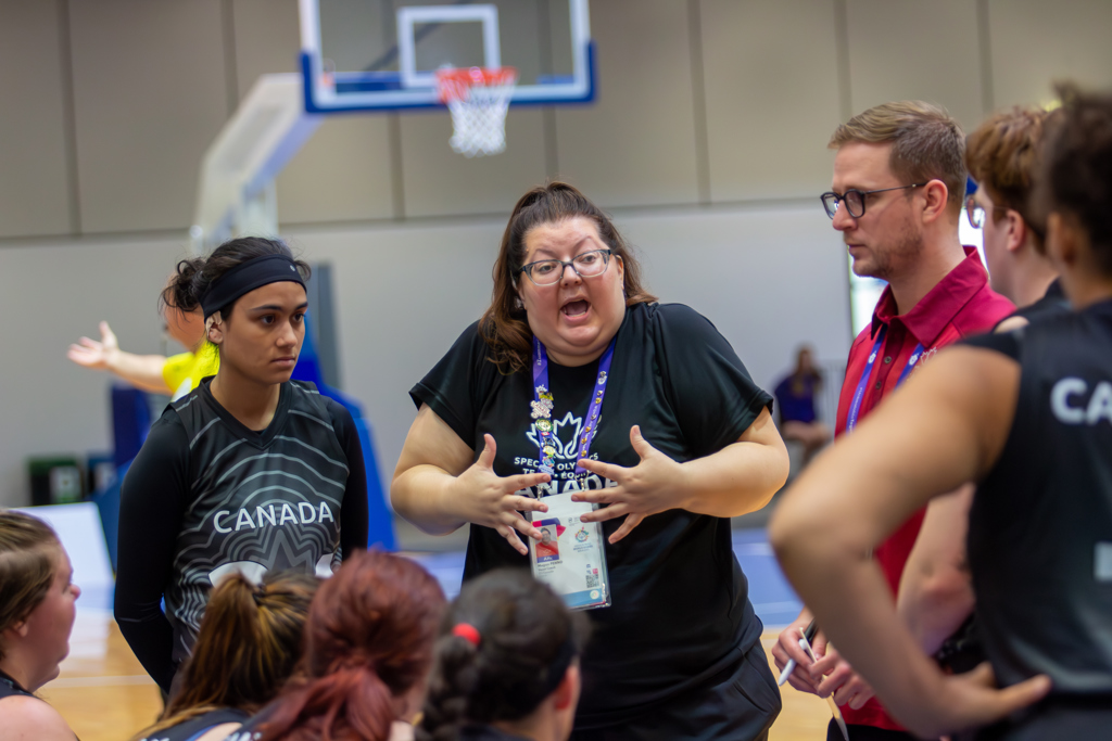 A woman volunteer is passionately speaking to the team. She is wearing a black Special Olympics Team Canada t-shirt with a blue lanyard around her neck. The team around her is listening intently to what she is saying.