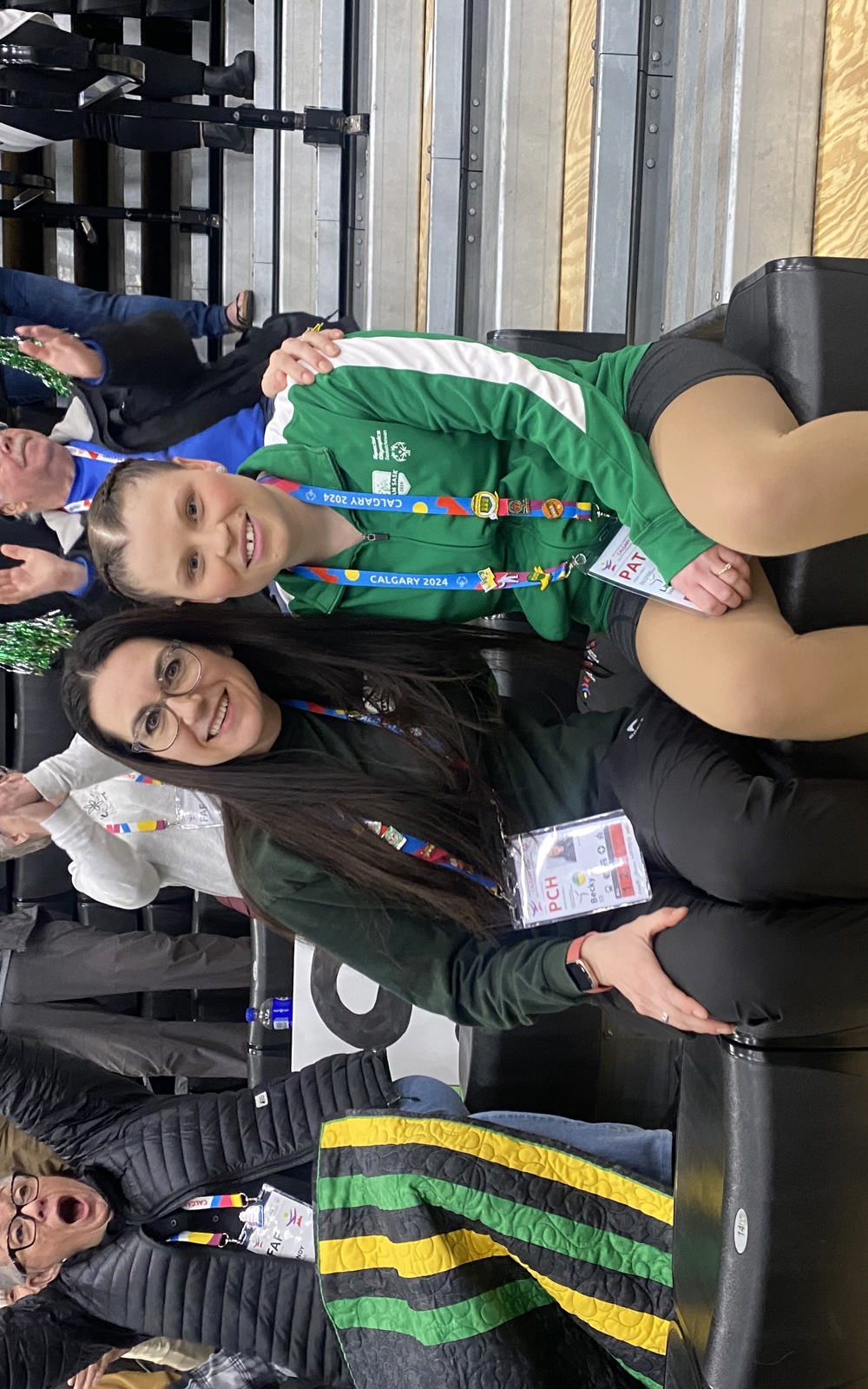 An athlete is sitting with her coach. Their arms are wrapped around behind each other. They are sitting in the bleachers of a sport event. The people behind them are all cheering.