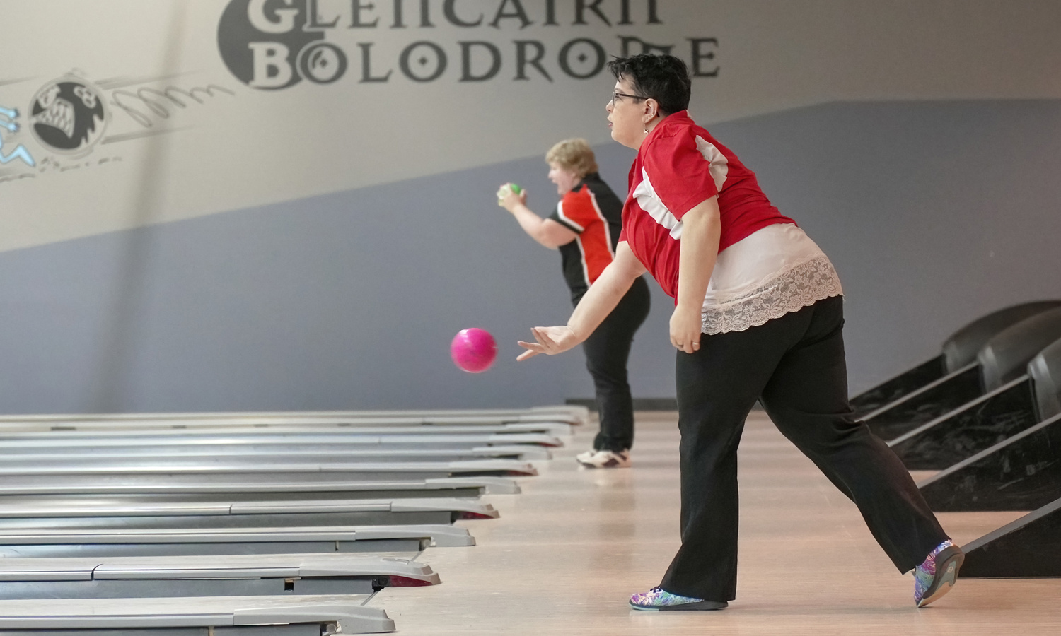 An action shot photo of a bowling athlete. The photo is taken from the side and you can see the start of many different bowling lanes. In the photo, two bowlers can be seen playing the game. The bowler closest to the camera is throwing a pink bowling ball which can be seen midair.