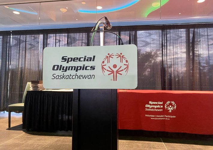 An image of a podium at a fancy event. The podium has a sign on it that reads, "Special Olympics Saskatchewan". Behind the podium is a long table in a red table cloth. The tablecloth also reads, "Special Olympics Saskatchewan".