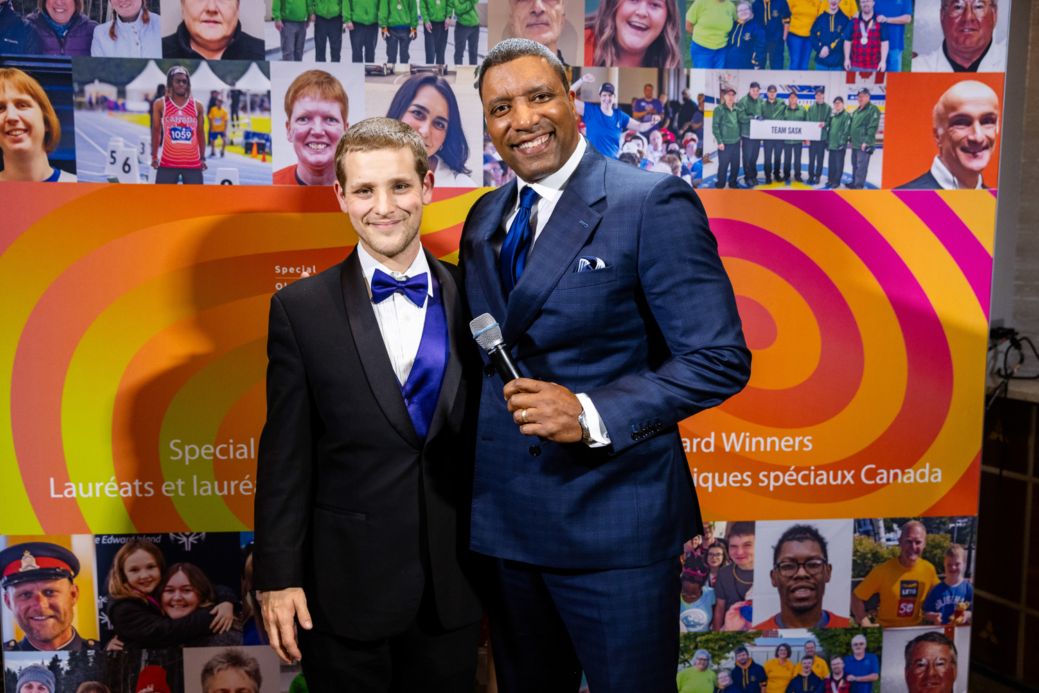 Two men in suits smiling at the camera. One has a microphone in his hand 