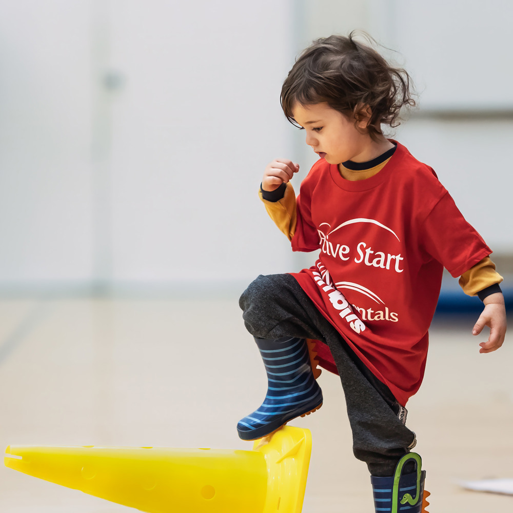 Little girl in a red active start shirt, blue stripped rainboots with her foot up on a yellow cone that is lying on its side. Background is a gymnasium.  