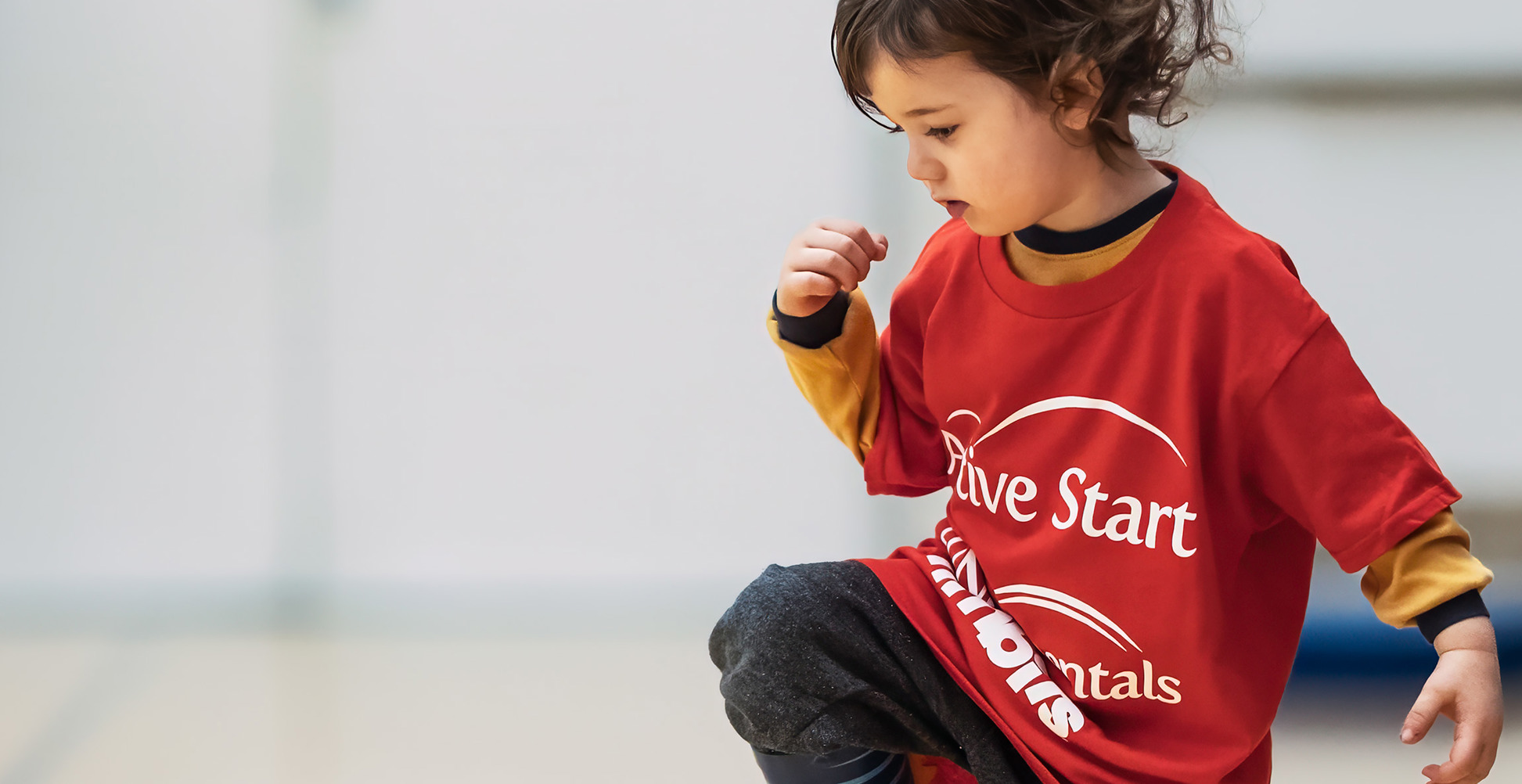 Little girl in a red active start shirt, blue stripped rainboots with her foot up on a yellow cone that is lying on its side. Background is a gymnasium.  