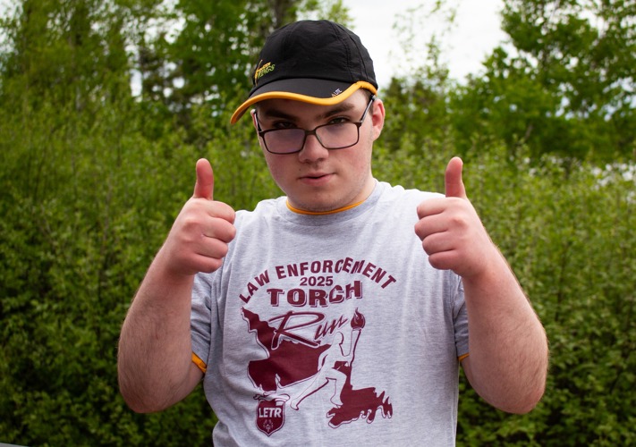 A half body photo of an athlete giving the camera two thumbs up. He is wearing a grey t-shirt that reads "Law Enforcement 2025 Torch Run" on it and a black baseball cap. Behind him are lots of greenery, trees, and bushes.