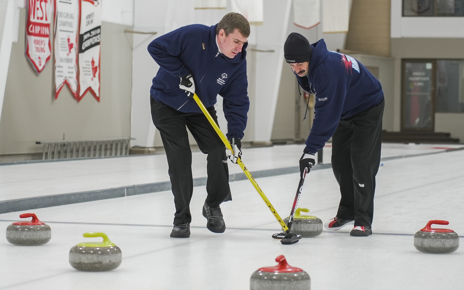 Two athletes participating in a curling match. There are three red and two yellow curling stones dispersed across the ice. The two athletes are using their curling sticks to allow one of the yellow curling stones to move easier.
