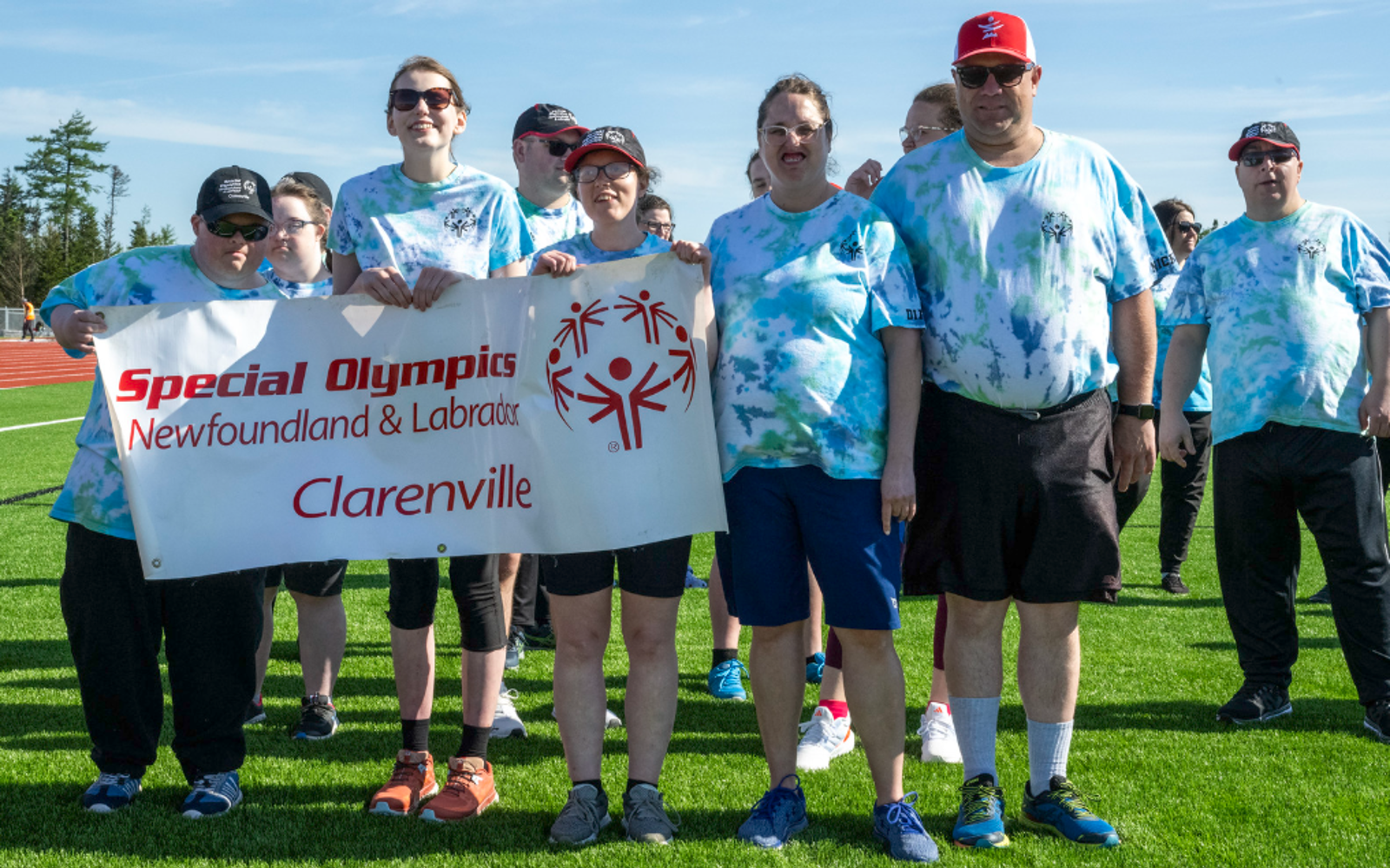 A group photo of the Clarenville Allstars Community Club. Everyone in the photo is wearing a blue tie-dye shirt. Three athletes are holding up a banner that says "Special Olympics Newfoundland & Labrador Clarenville".