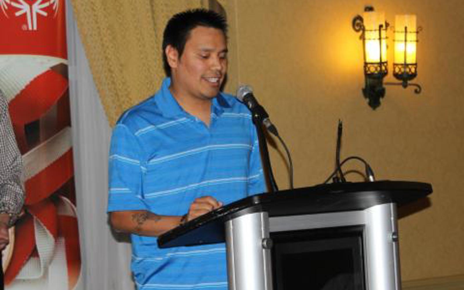 An athlete in a blue striped t-shit is standing at a podium giving a speech.
