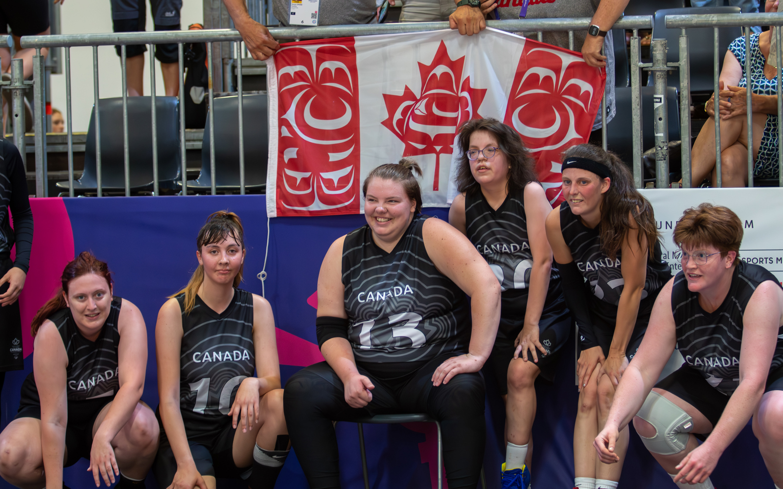 Six Special Olympics Saskatchewan athletes are sitting on the sidelines waiting for their turn to play. They are wearing black Canada jerseys with a different number on each of them. Supporters in the stands behind them are holding a Canadian flag.
