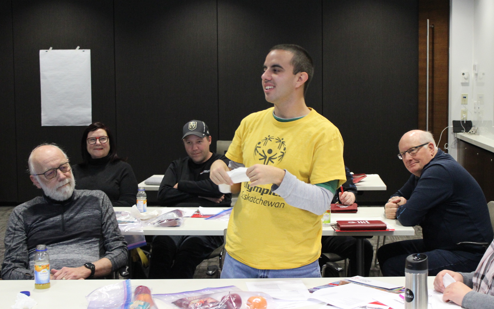 A conference room with three rectangular tables. People can be seen sitting at these three tables. One individual, in the middle of the image, in a yellow shirt, is standing up and presenting something to his peers. Everyone around him is listening intently.