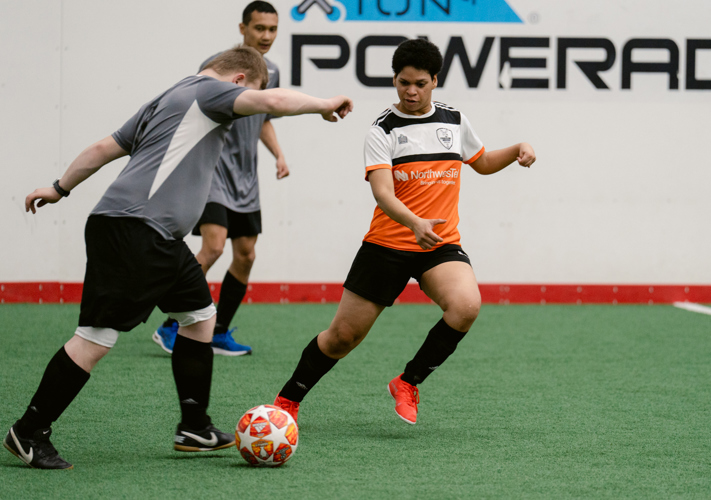 Three athletes playing soccer in an indoor soccer field. One athlete is wearing an orange jersey while the other two athletes are both in grey jerseys.