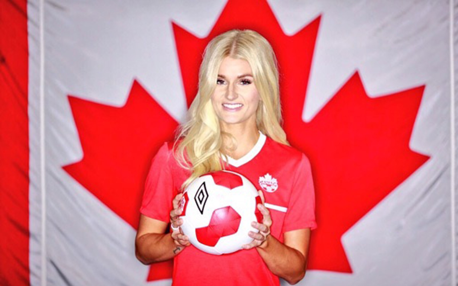 Headshot of a woman with long blond hair in front of a Canadian flag holding a soccer ball.  