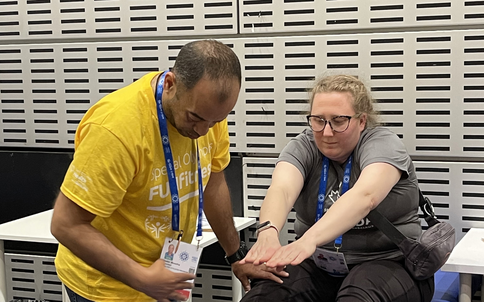 Young blond woman with glasses sitting on a blue medical bed. She is wearing dark pants and a grey shirt and stretching to reach her toes. A man in a yellow shirt is beside her bending down to check her flexibility. 
