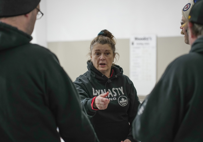 A coach, coaching her team. She is in the middle of speaking and pointing at an athlete. Two athletes can be seen in front of her, but they have their backs to the camera and are out of focus.