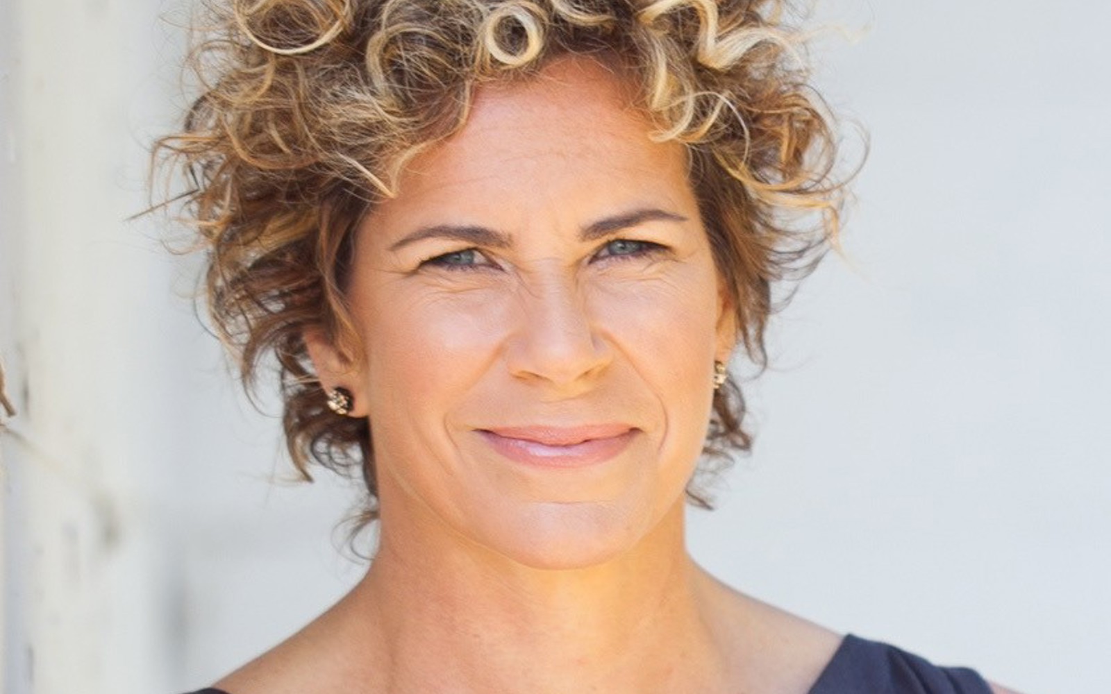 Headshot of a woman with curly blond short hair wearing a blue sleeveless blouse. 