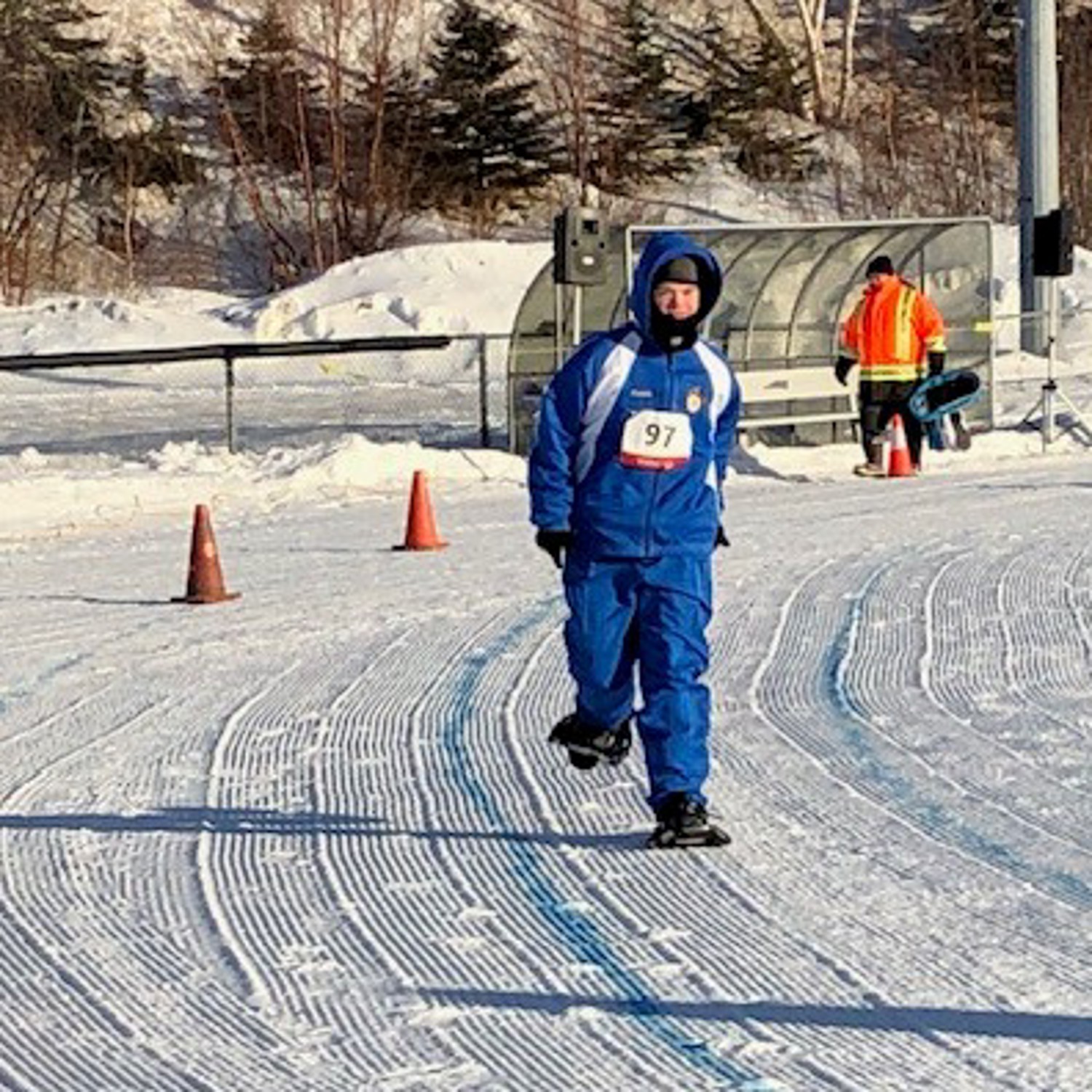 A photo of a snowshoeing athlete participating in his match. He is running around a curve that has been outlined with orange pylons. It is the winter and the athlete is wearing a blue snowsuit with the number "97" pinned to his coat.