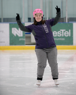 A figure skating athlete is posing for the camera. She is standing on the ice in a purple Yukon sweater and pink helmet, and has her arms up showing peace signs. She is smiling and happy.