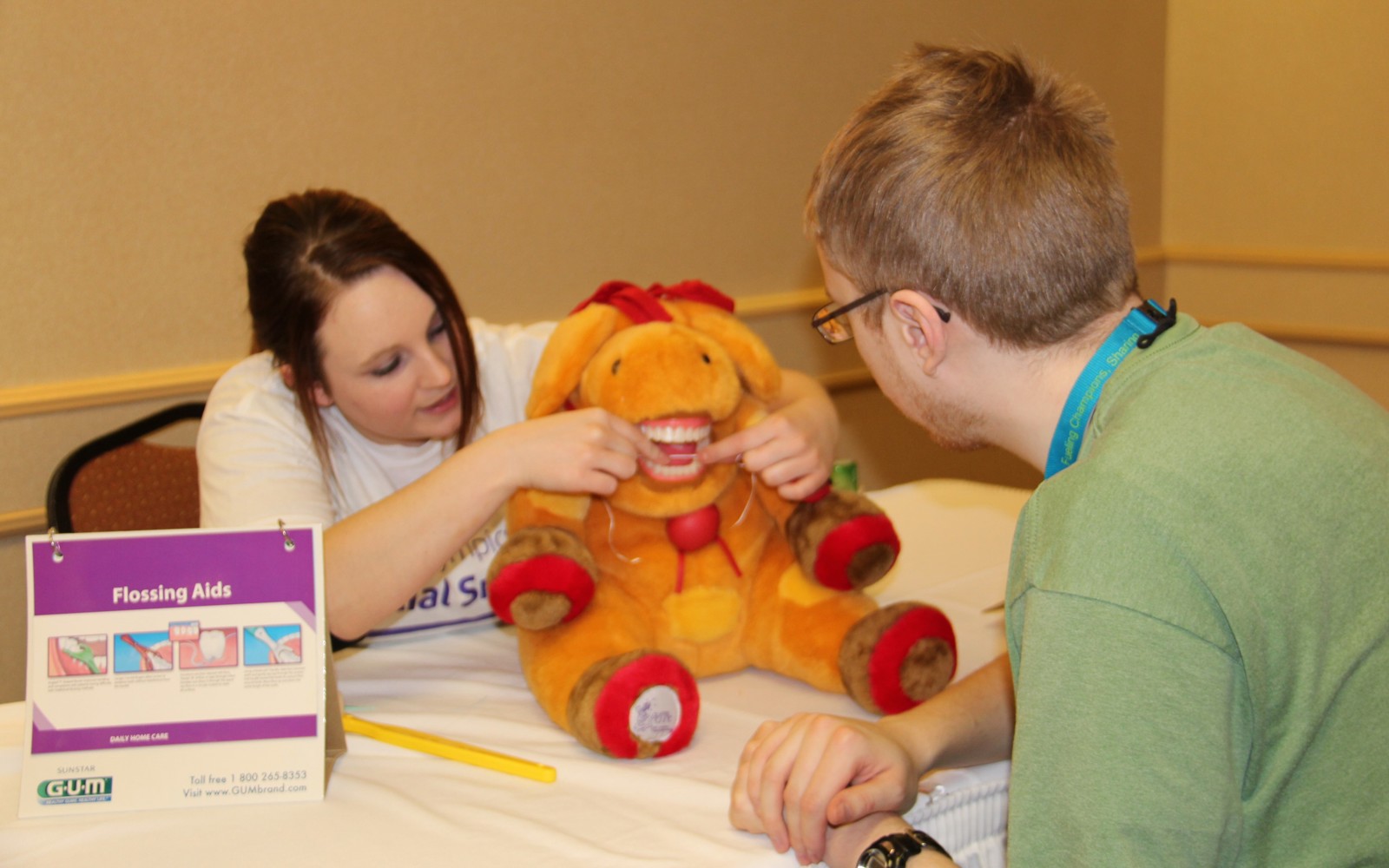 A Healthy Athletes practitioner is demonstrating how to floss your teeth on a stuffed animal with teeth. A athlete in a green t-shirt can be seen from behind watching the demonstration.