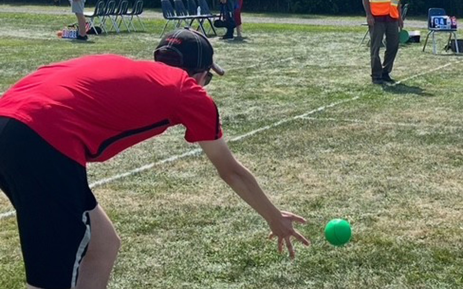 Man in black shorts and red shirt throwing a bocce ball on green grass 