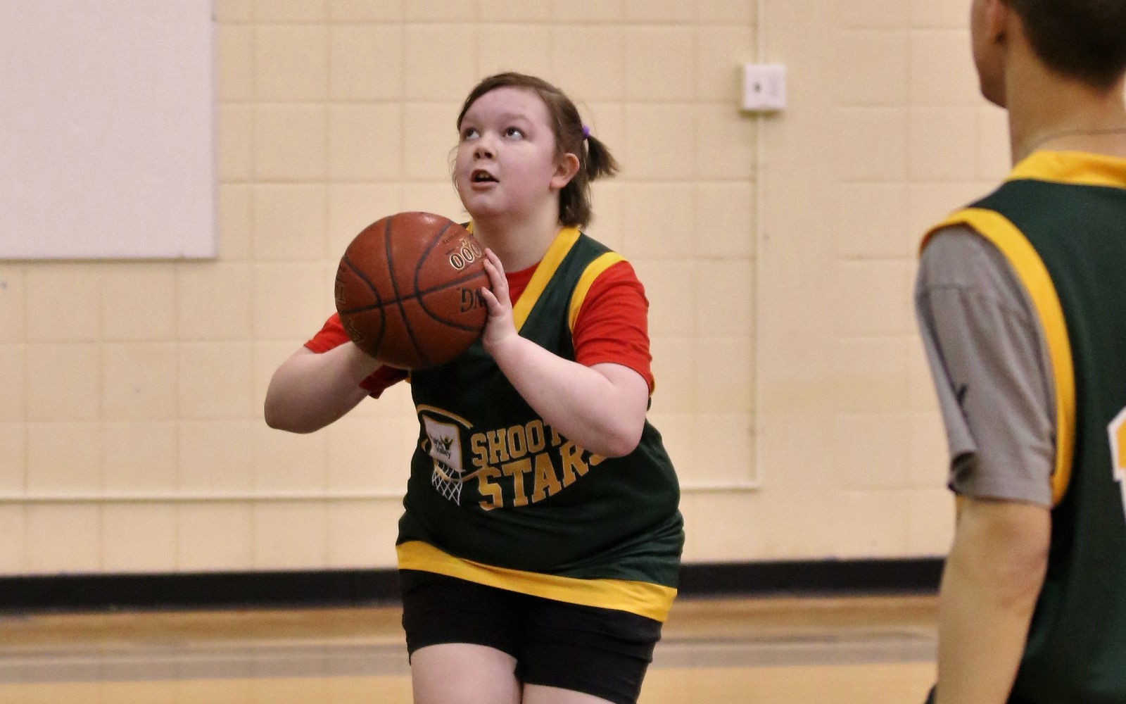 Two basketball athletes can be seen on the court. One athlete, you can only see their back. The other athlete is looking up and about to throw the ball in the hoop.
