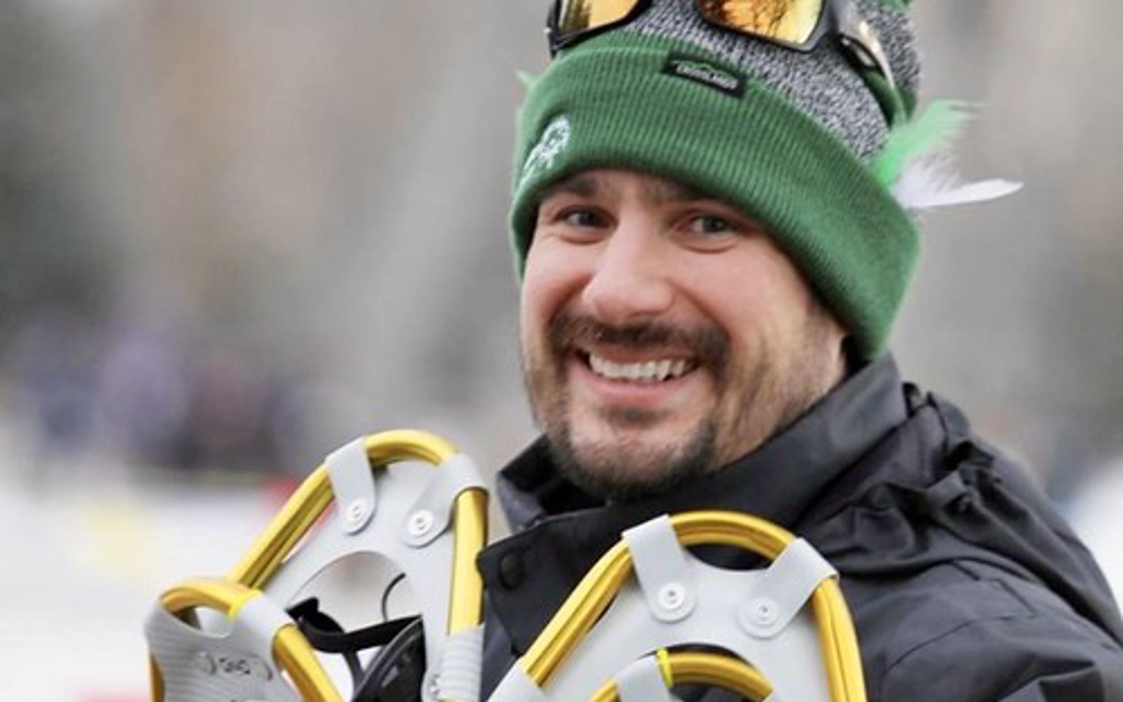 A close up image of Eric, a Special Olympics Saskatchewan coach. He is smiling for the camera and holding snowshoes in his hands. The background behind him is blurred.