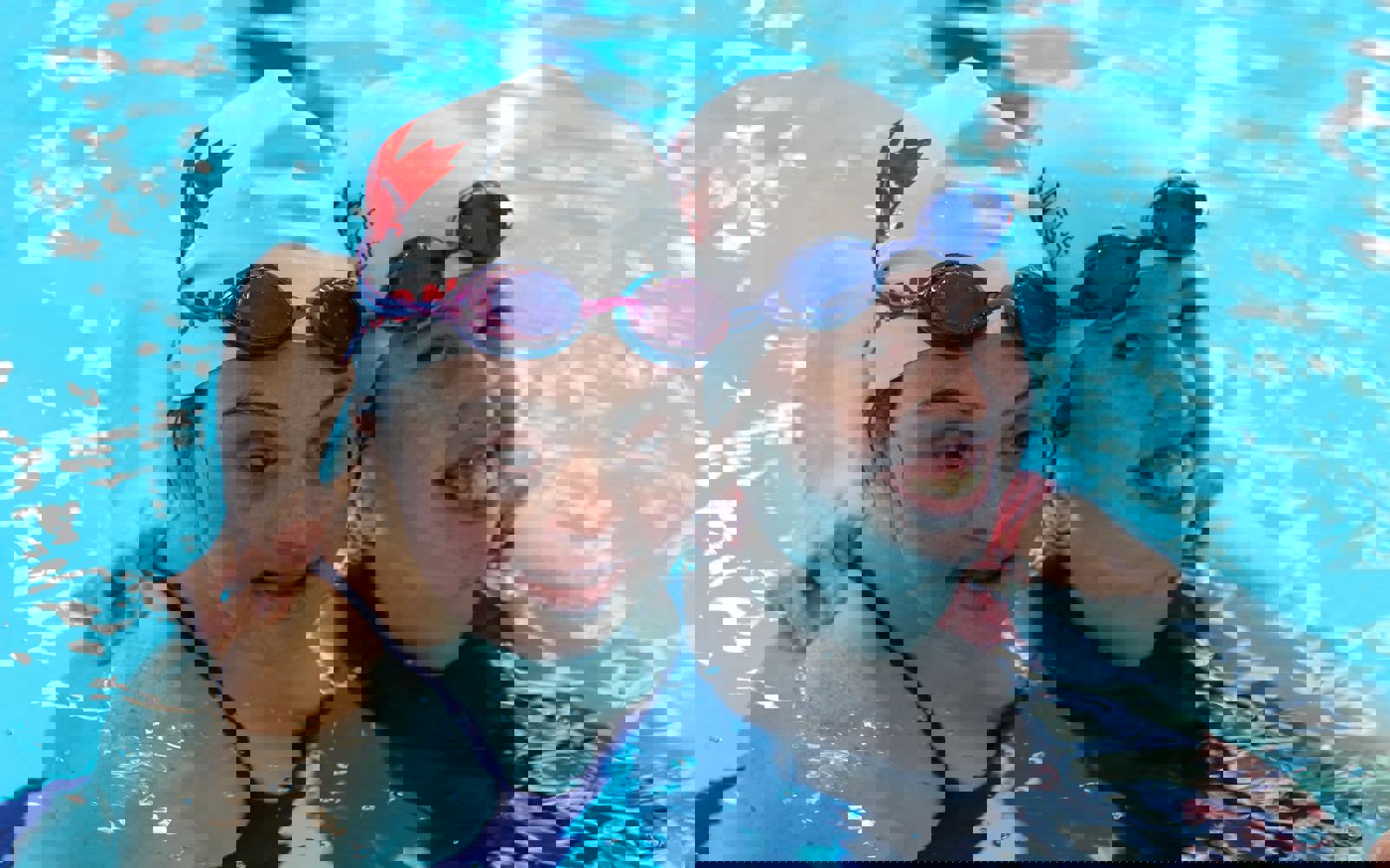 Two girls in a swimming pool. Both are wearing swim caps and have googles on their heads. One is smiling at the camera and the other is looking down at the pool.  