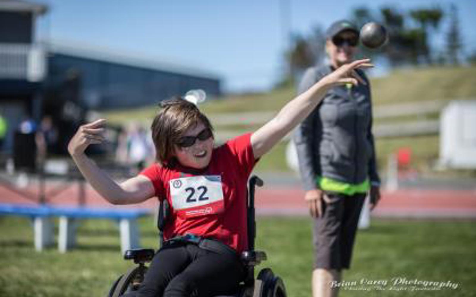 Person in a wheelchair with a red shirt and a number tag throwing a shot put 