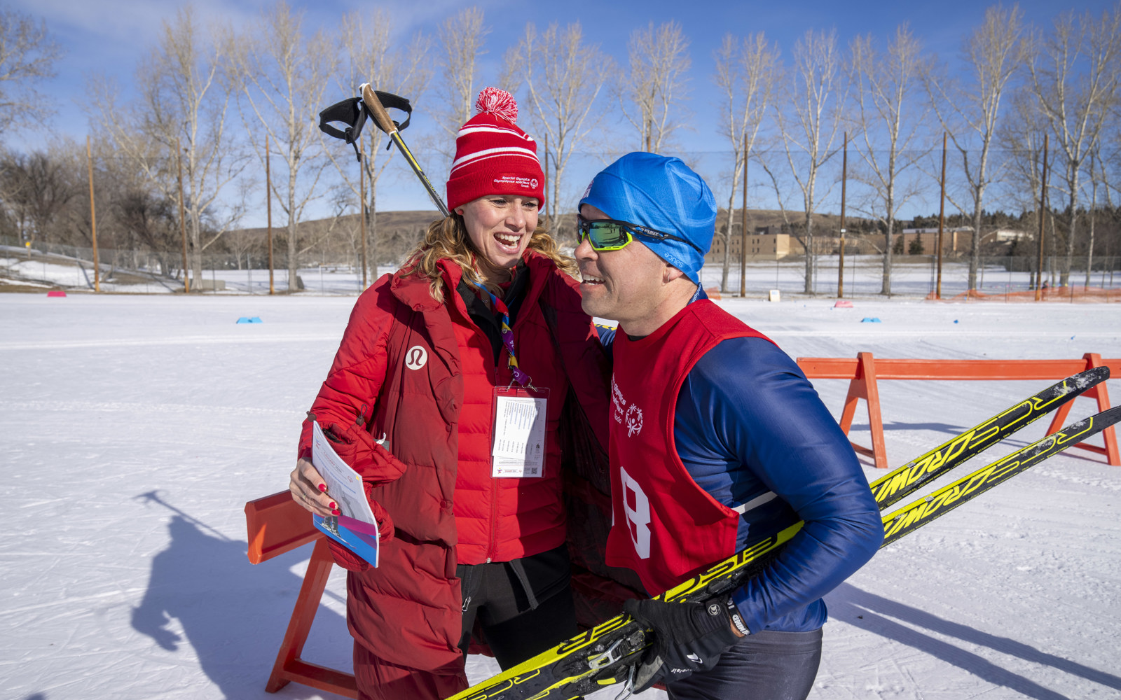 Man in a blue skit suit with a red pinnie and a blue hat wearing goggle.s. He is holding yellow skis and smiling. A women in a red coat and red hat with blond hair stands beside him smiling. 