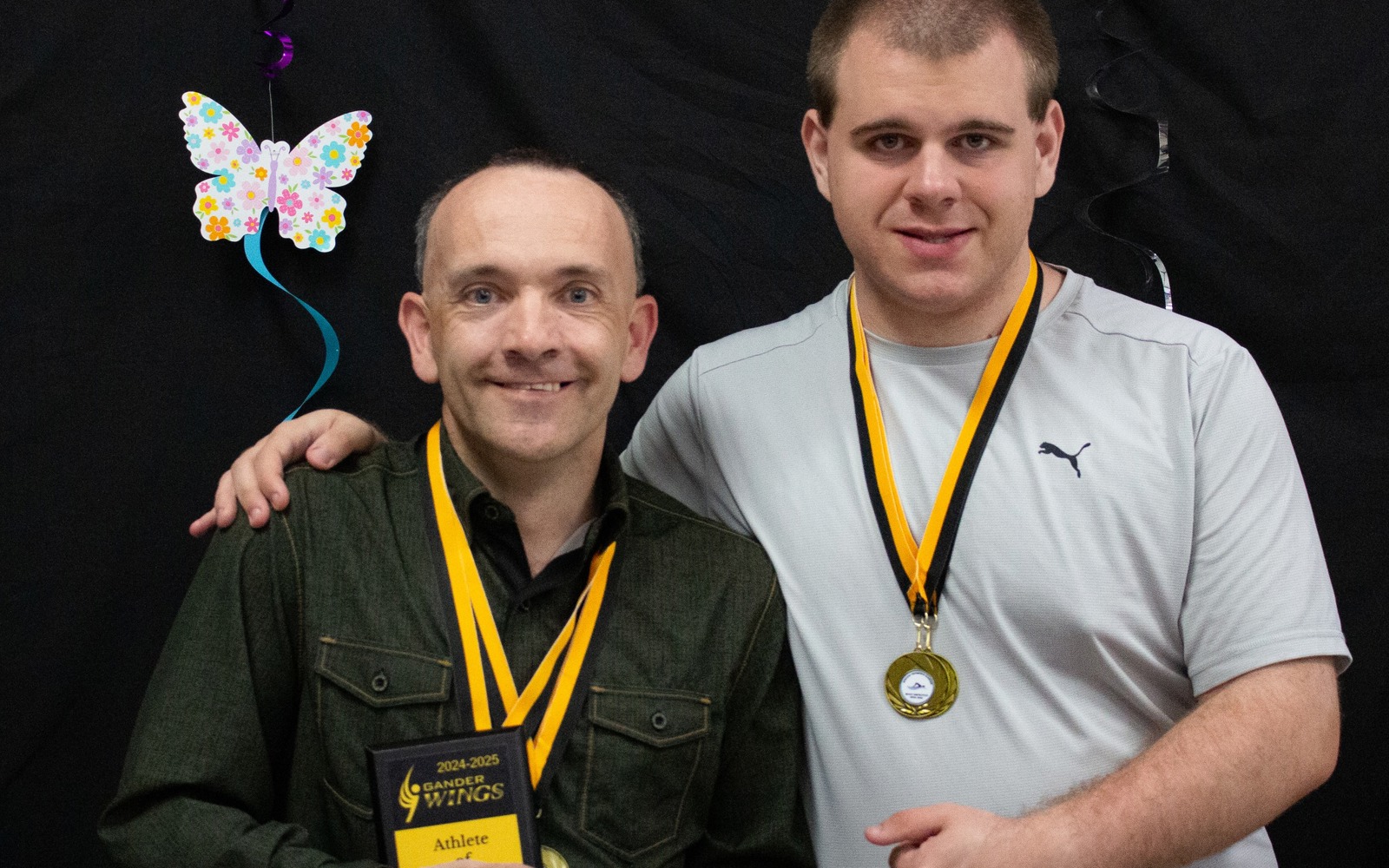 Two Special Olympics Newfoundland & Labrador athletes are smiling and posing for the camera. They both have medals hanging around their necks, and one athlete on the left is holding a plaque he won. The athlete on the right has one arm around the athlete on the left and his holding his other hand with the athlete.