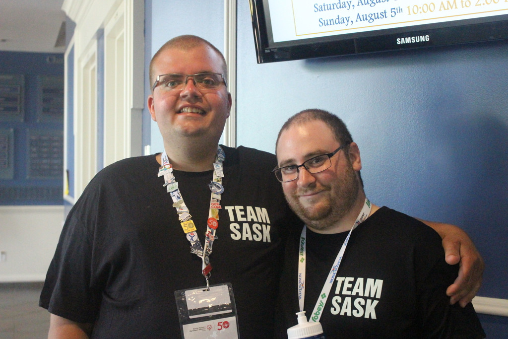 Two Special Olympics Saskatchewan athletes have their arms around and behind each other and are smiling for the camera. They are both wearing black "Team Sask" t-shirts and colourful lanyards.
