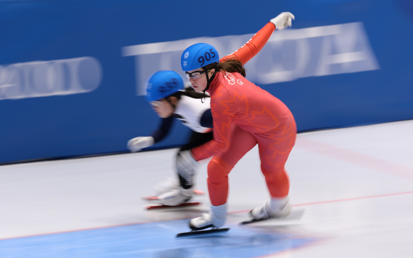 Canadian female speed skater in red skin suit. In the background is another speed skater in a black and white skin suit. 