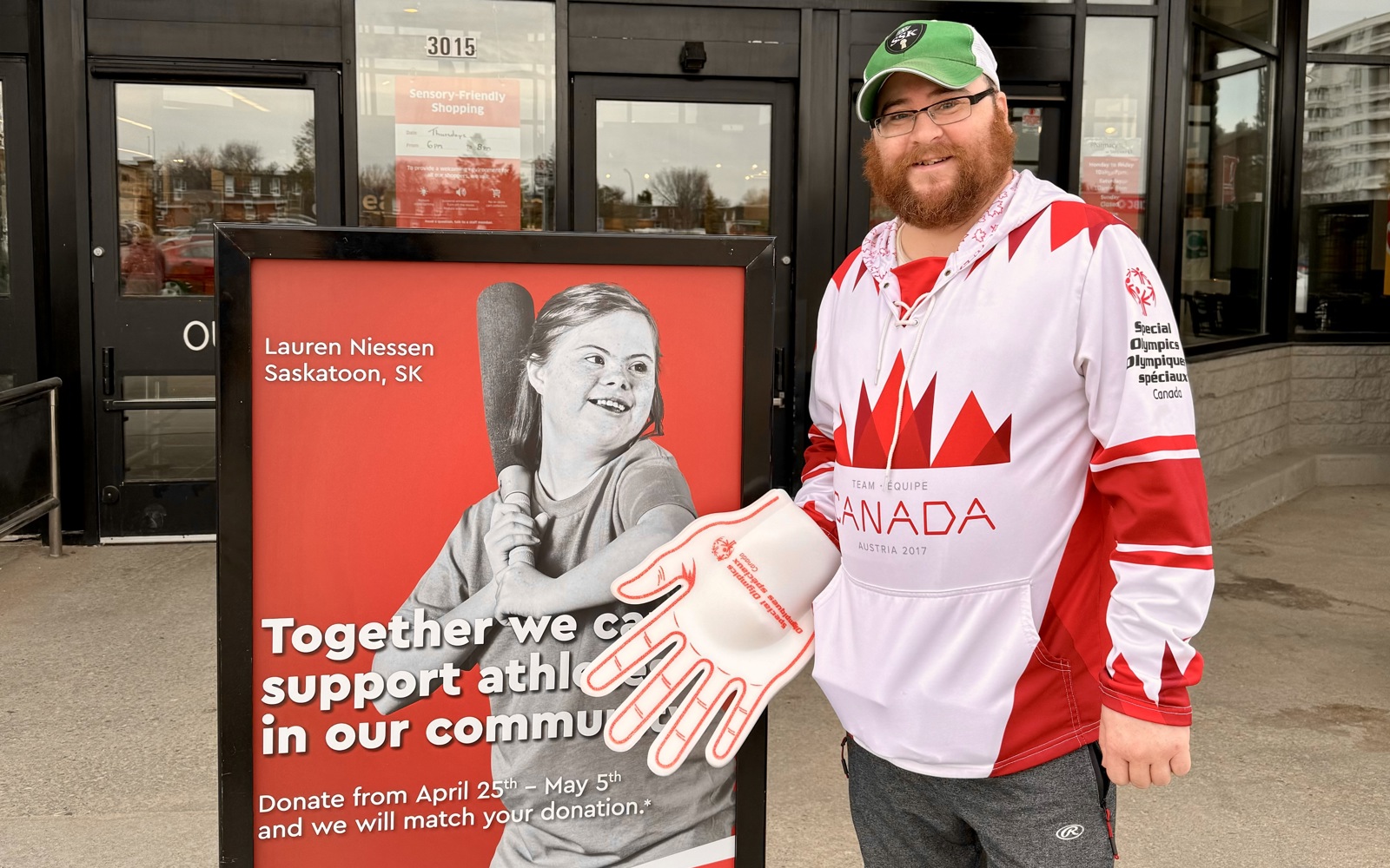 A male athlete with a red, Canada, Special Olympics Saskatchewan jersey, a green baseball cap, and a red and white foam hand, poses and smiles for the camera in front of a donation sign.