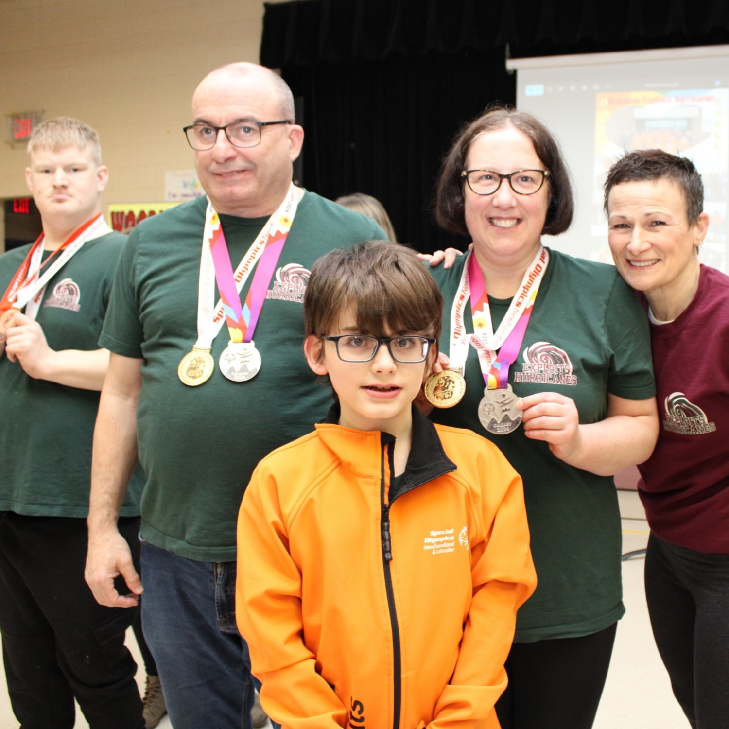 A group photo of multiple people. In the front is a young athlete wearing an orange Special Olympics Newfoundland & Labrador sweater. Behind him are three Special Olympics Newfoundland & Labrador athletes with medals hanging around their necks, as well as a volunteer.