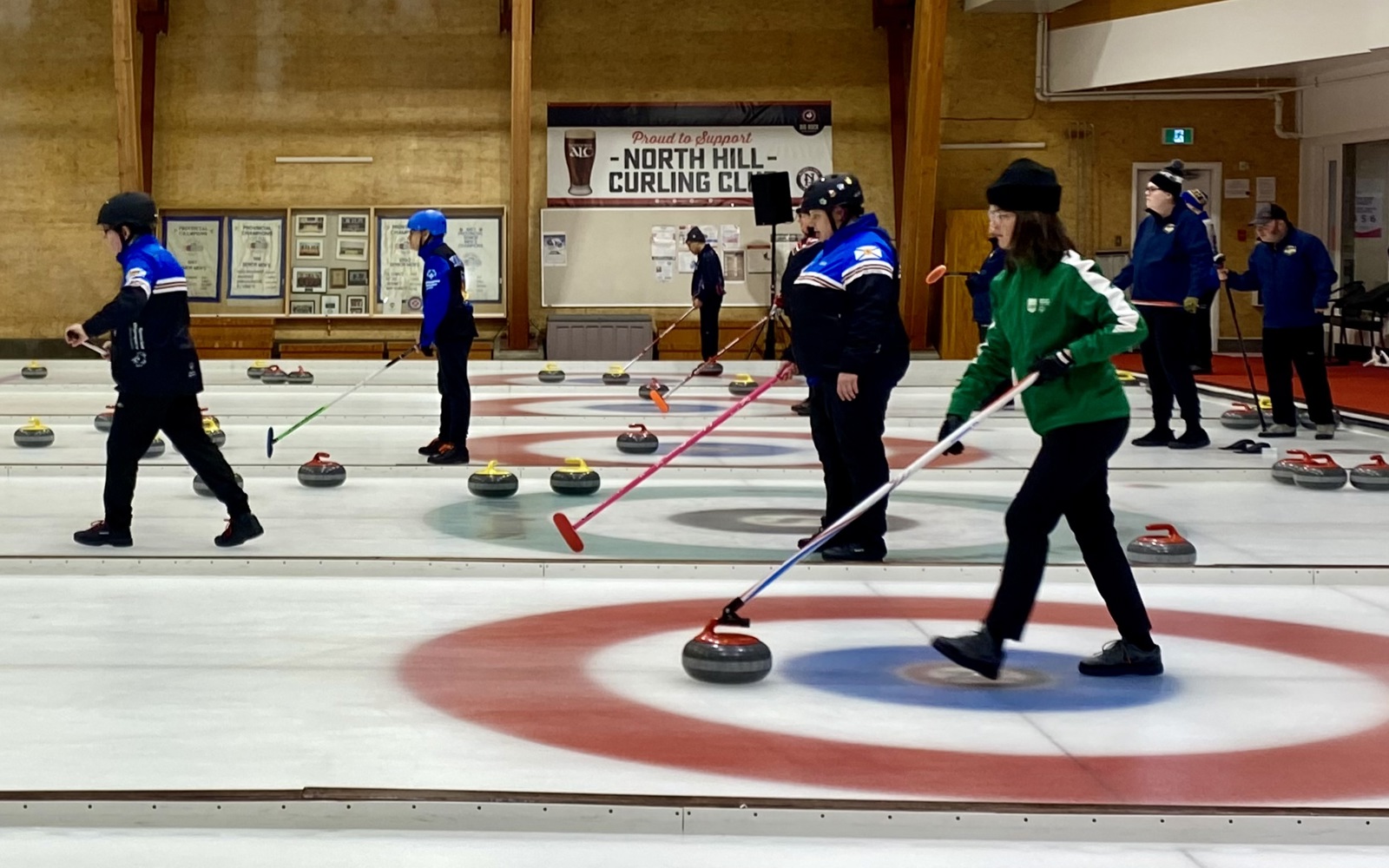A wide shot action photo of curling games in play. Multiple curling athletes can be seen with their curling sticks, getting ready to move the curling stone across the ice.