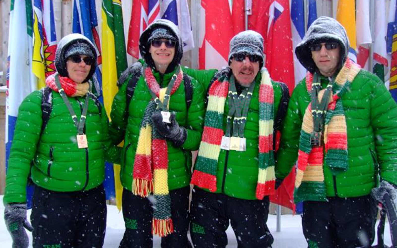 Four athletes are posing for a group photo in matching green puffer jackets and green, yellow, and red striped scarves. They are smiling for the camera as snow is falling around them. Behind them, are different flags.