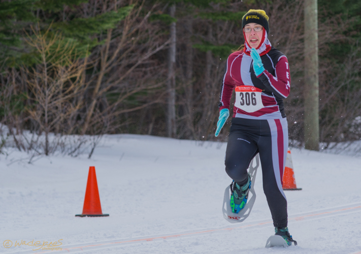 An action shot photo of a snowshoeing athlete in a competition. She is running down the track that is lined with orange pylons. She is dressed in athleisure fit for her sport and the winter weather, and has the number "306" pinned to her top.