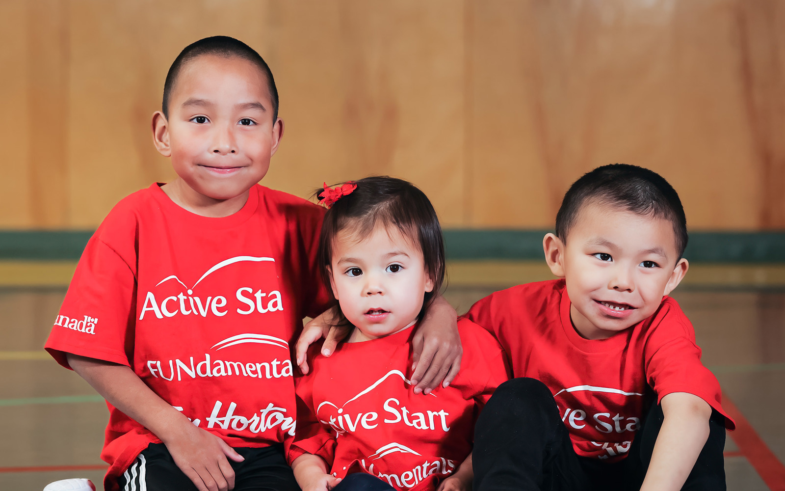 Three young people - two boys and a girl - sitting on the floor. They appear to be about 5-7 years old. All are wearing red shirts that say ACTIVE START. All of them are smiling. 