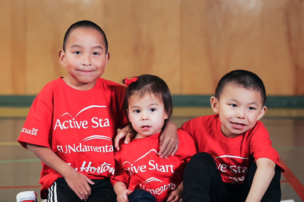 Three young people - two boys and a girl - sitting on the floor. They appear to be about 5-7 years old. All are wearing red shirts that say ACTIVE START. All of them are smiling. 