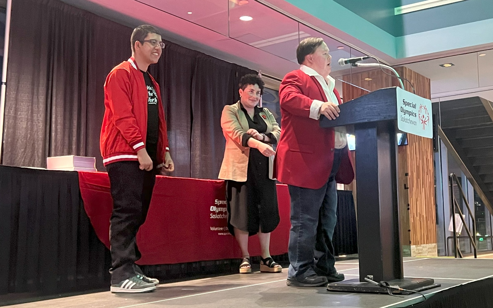 Three individuals are standing on a stage. The angle is taken from off stage looking up at them diagonally. Two individuals are standing back while one is at the podium giving a speech.