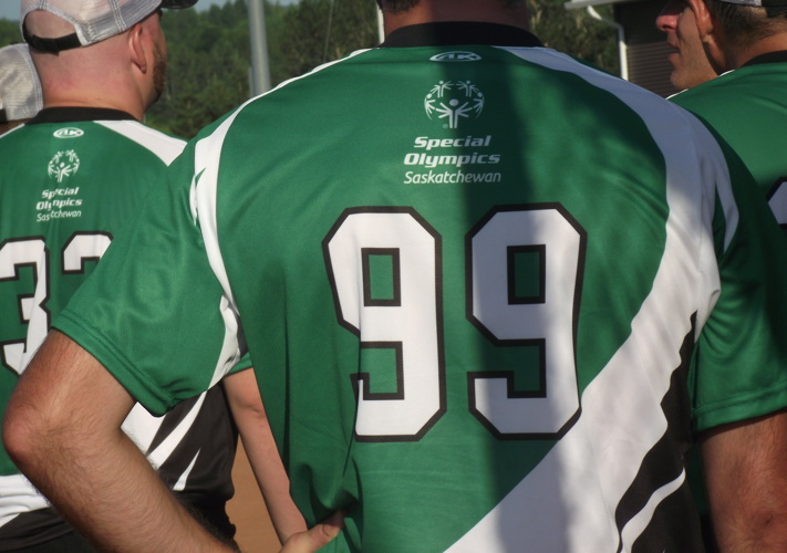 Multiple athletes are grouped together. We can only see their backs as the image is a close up of the back of an athlete's jersey. The jersey is green and has the Special Olympics Saskatchewan logo and the number 99 on it.