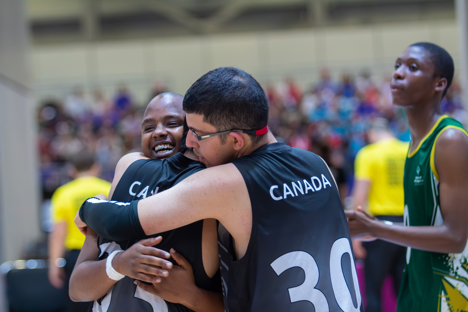 Three athletes engaging in a group hug. For two of the athletes we can only see their backs and they are wearing black "Canada" jerseys. The one athlete we can see is smiling very happily.