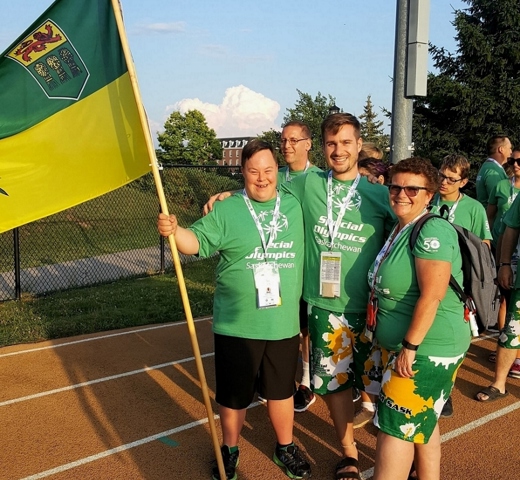 Three Special Olympics Saskatchewan athletes in green "Special Olympics Saskatchewan" t-shirts, are standing on the track. They have their arms wrapped behind each other and are smiling for the camera. One athlete, the one on left, is holding a big Saskatchewan flag.