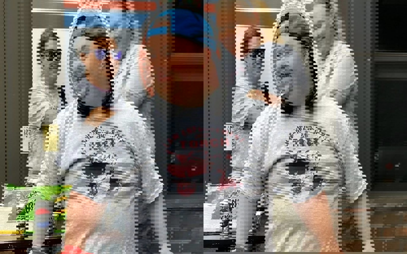 An athlete is walking towards the camera, but looking to the left. He is smiling happily with a hot dog in one hand and his water bottle in another. He is wearing a grey Law Enforcement Torch Run t-shirt and a blue baseball cap, facing backwards.