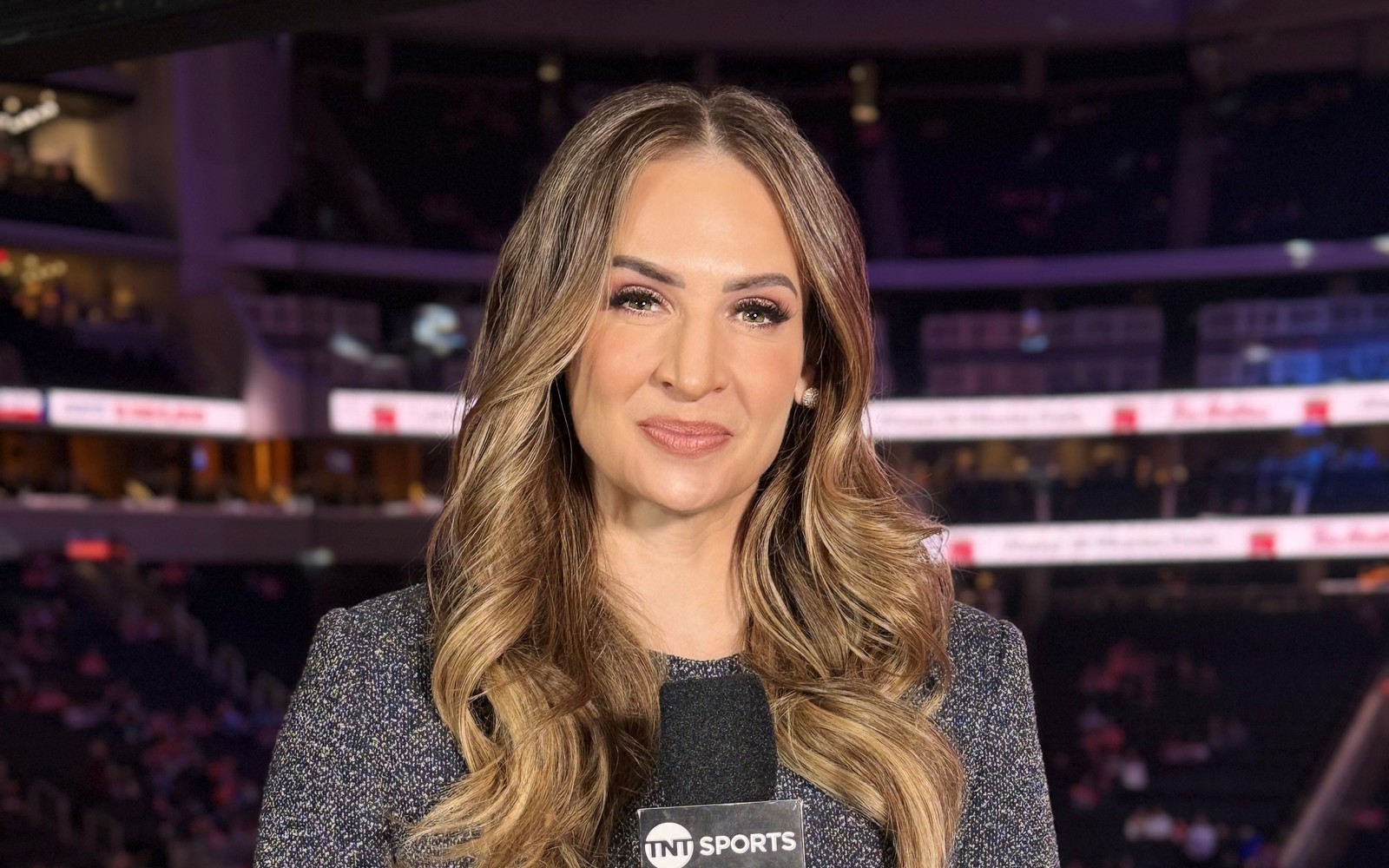 Headshot of a woman with curly long hair wearing a grey dress, holding a microphoe and standing in a hockey arena. 