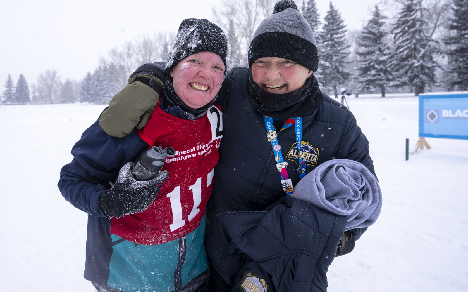 Two people with their arms around each other, smiling into the camera. One is a woman in a red pinny and one is a man with a black jacket. The background is covered in snow. 