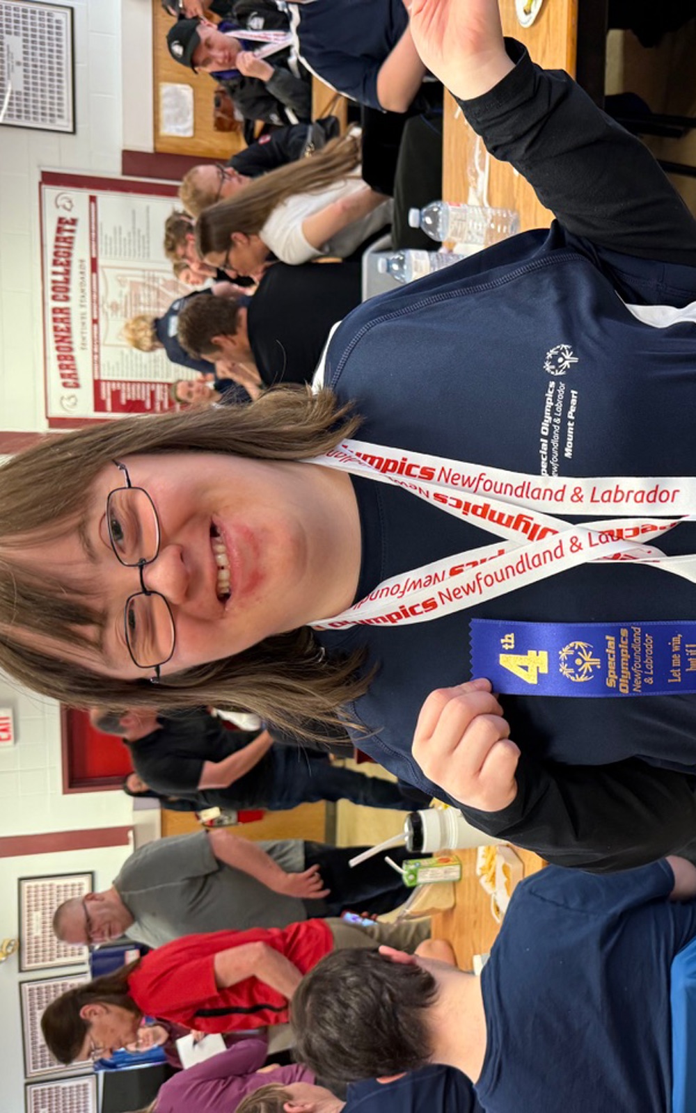 A Special Olympics Newfoundland & Labrador athlete is smiling for the camera and posing with her awards. She has two bronze medals hanging around her neck and she's holding a blue 4th place ribbon.